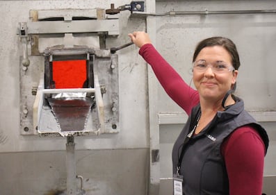 Janel Rowbotham stands next to a little door, showing bright red molten aluminum inside.